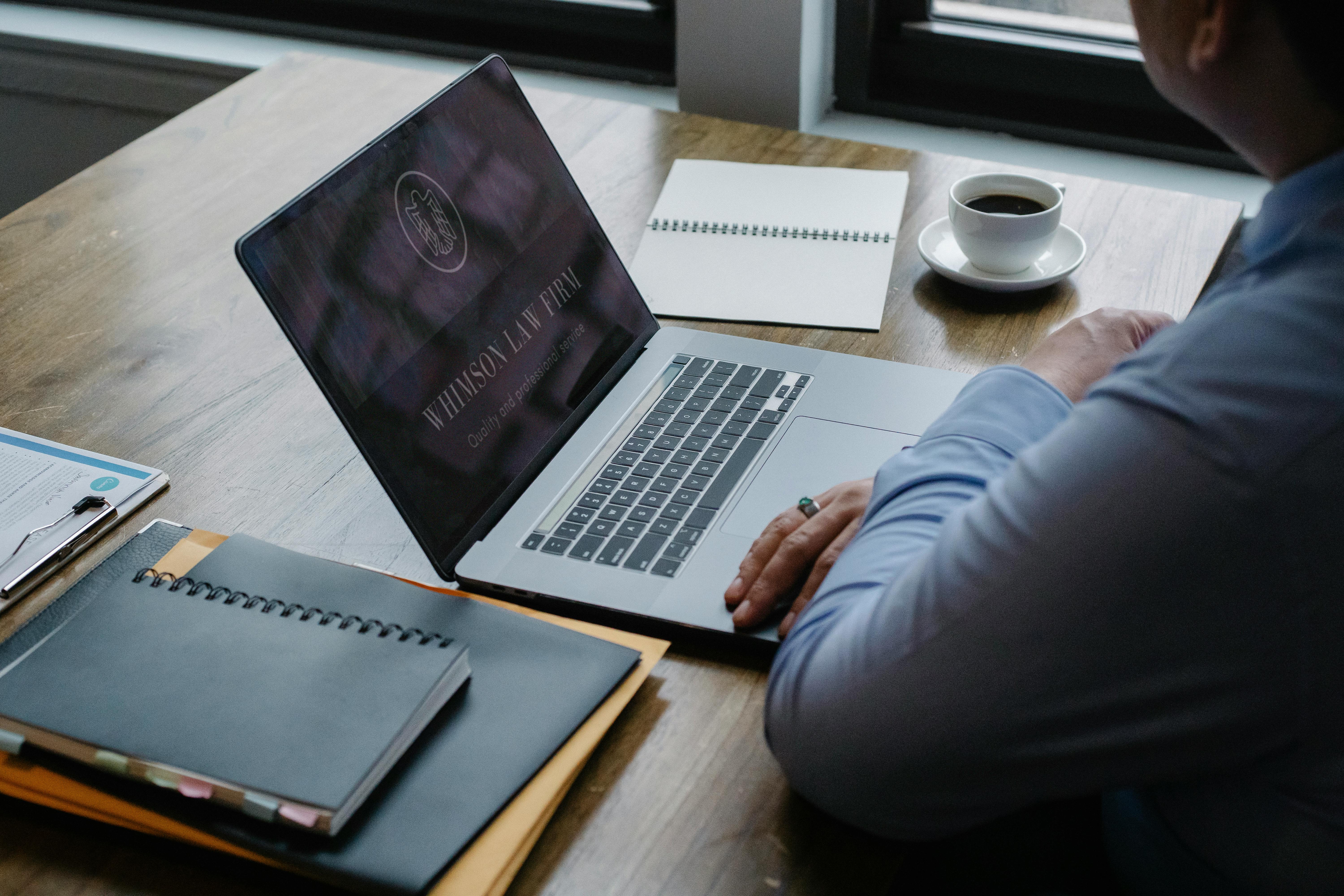 A man in shirtsleeves sitting at a desk with his laptop open to a law firm website and a small cup of coffee, a notebook, a planner, and some files on either side of it.