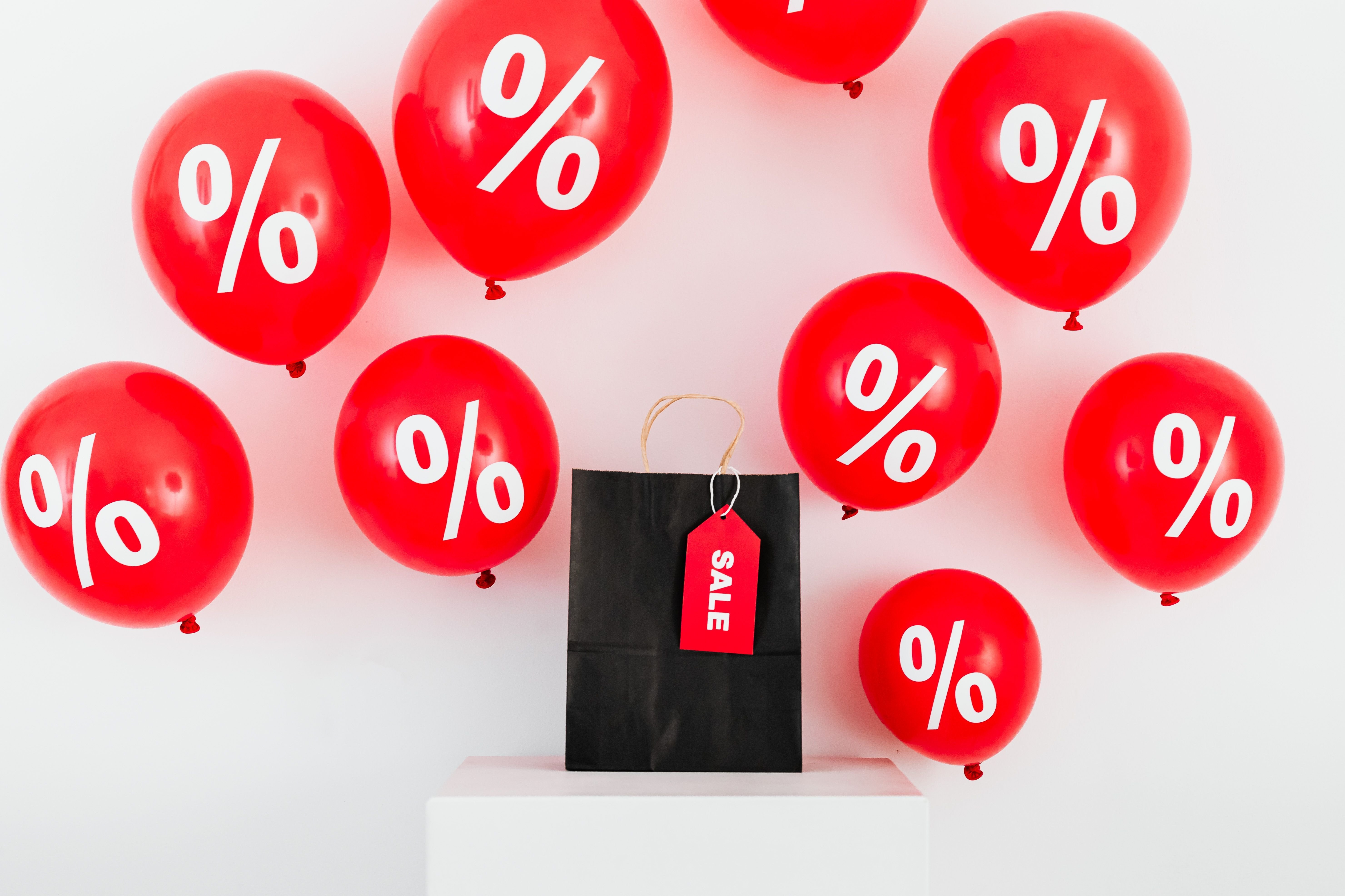 A gift bag with a red "Sale" tag hanging from one of its straps sits on a white pedestal in front of a white background with nine red balloons with white "%" signs floating around it.