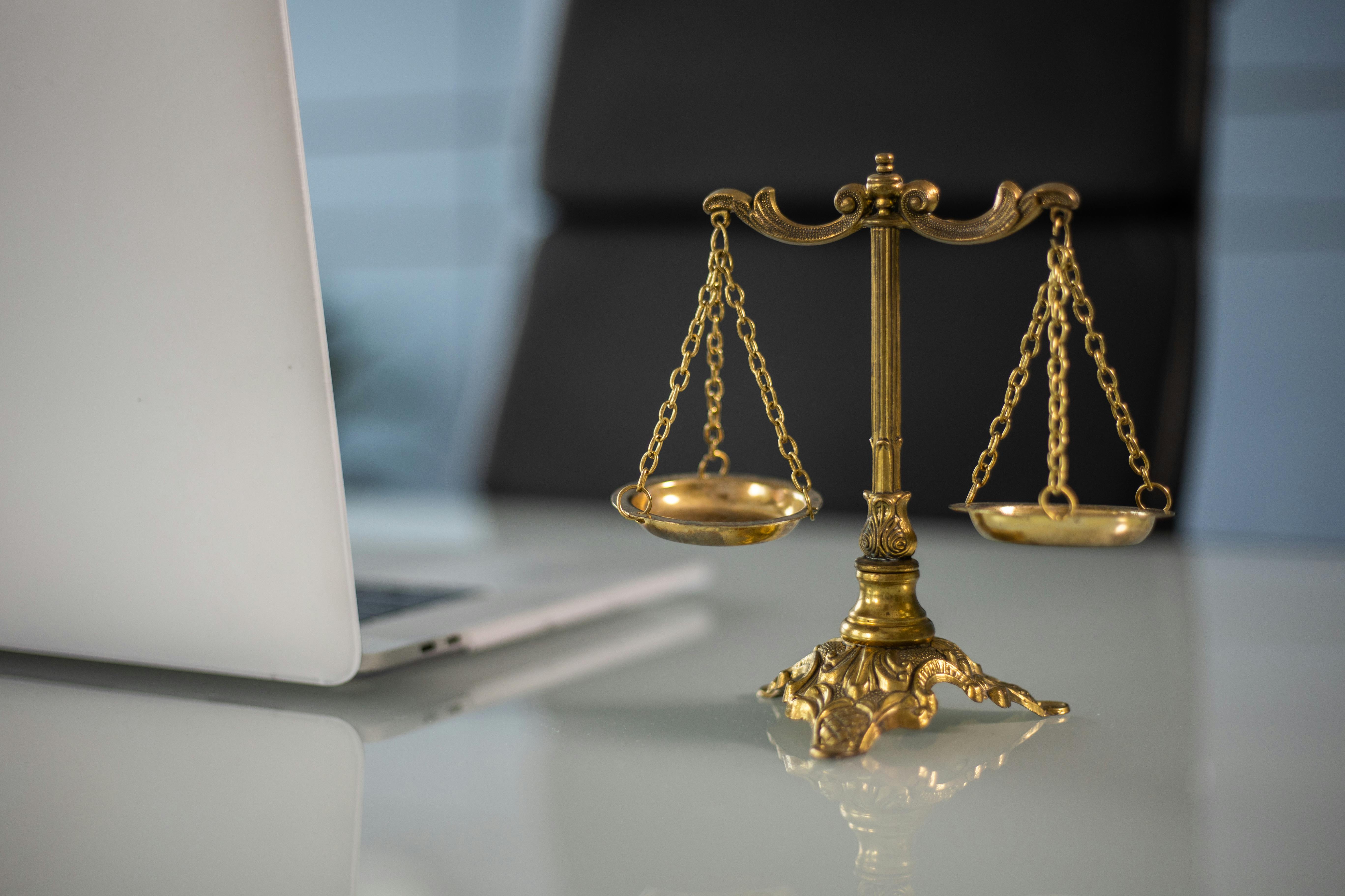 A small gold ornamental paperweight modeled after the scales of justice sits on a desk next to an open laptop computer.
