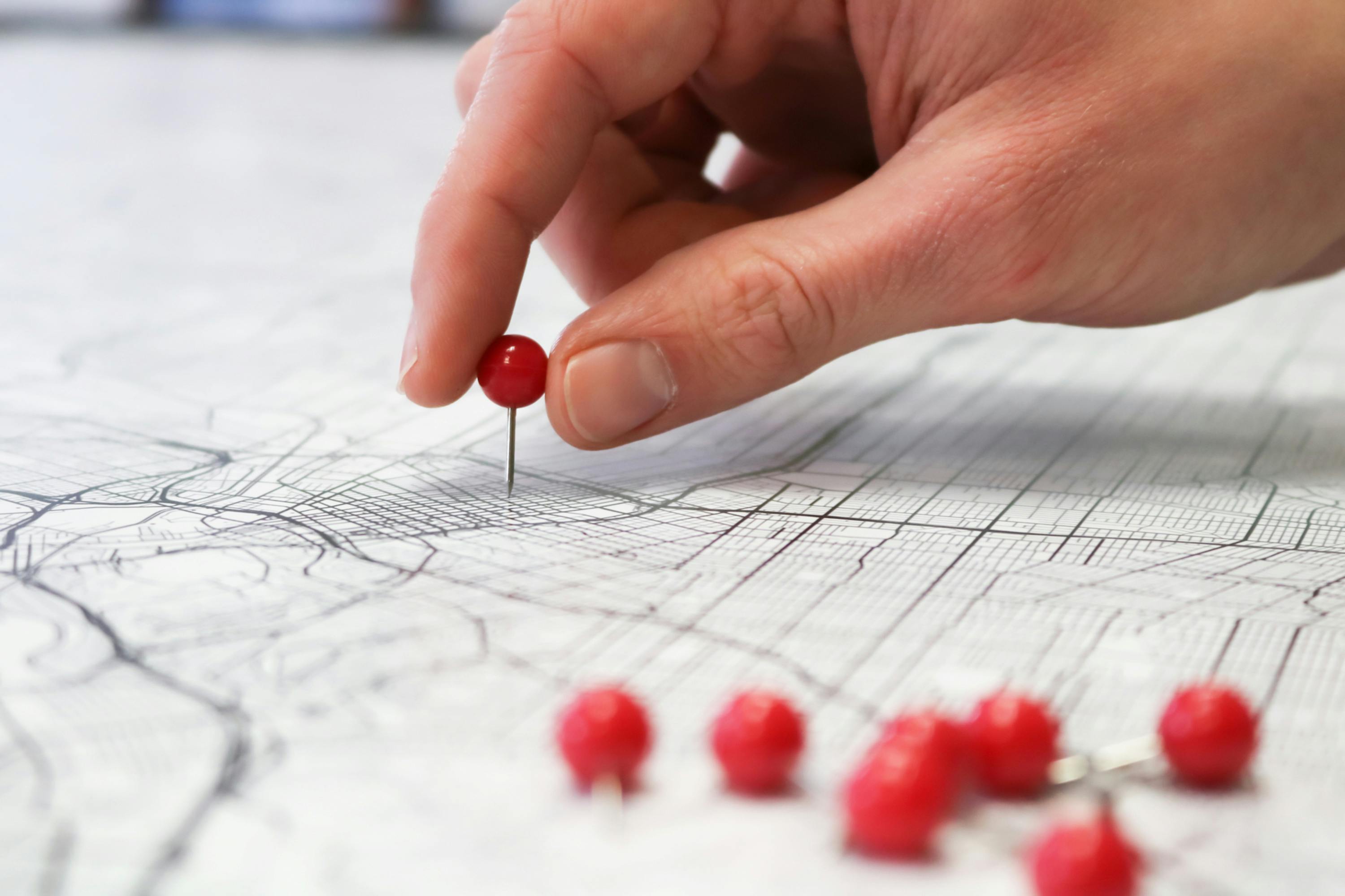 A hand putting a red push pin in a black and white paper map that is sprawled out across a table to mark a specific place.