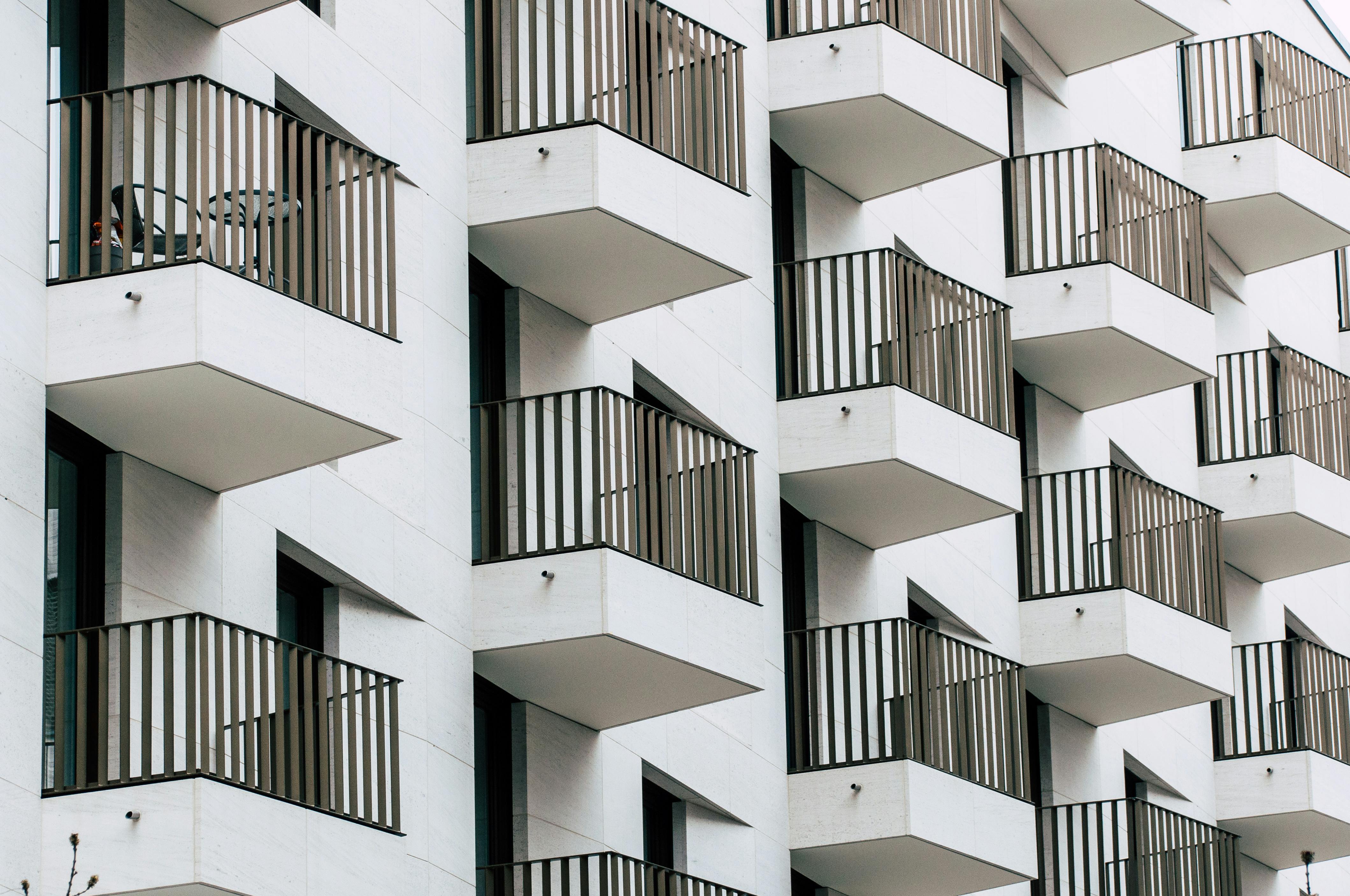 A white building with dozens of tiny balconies with black railings adorning it. The top balcony in the foreground has a small table and chair on it, but the rest appear empty.