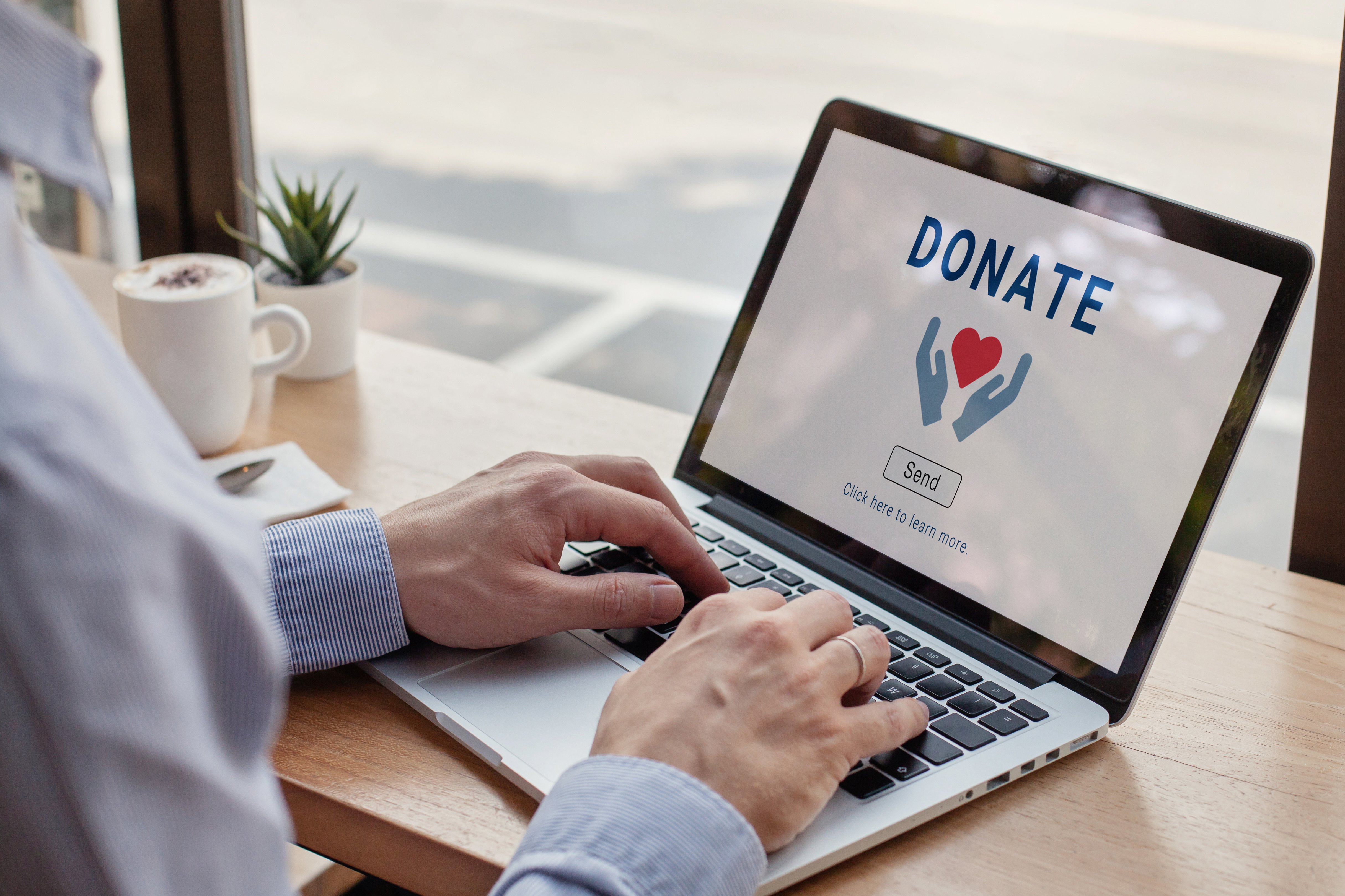 A man in shirtsleeves sitting at a desk typing on a laptop whose screen is on the Donations page for a nonprofit website, with "Donate" written in large letters at the top of the page.