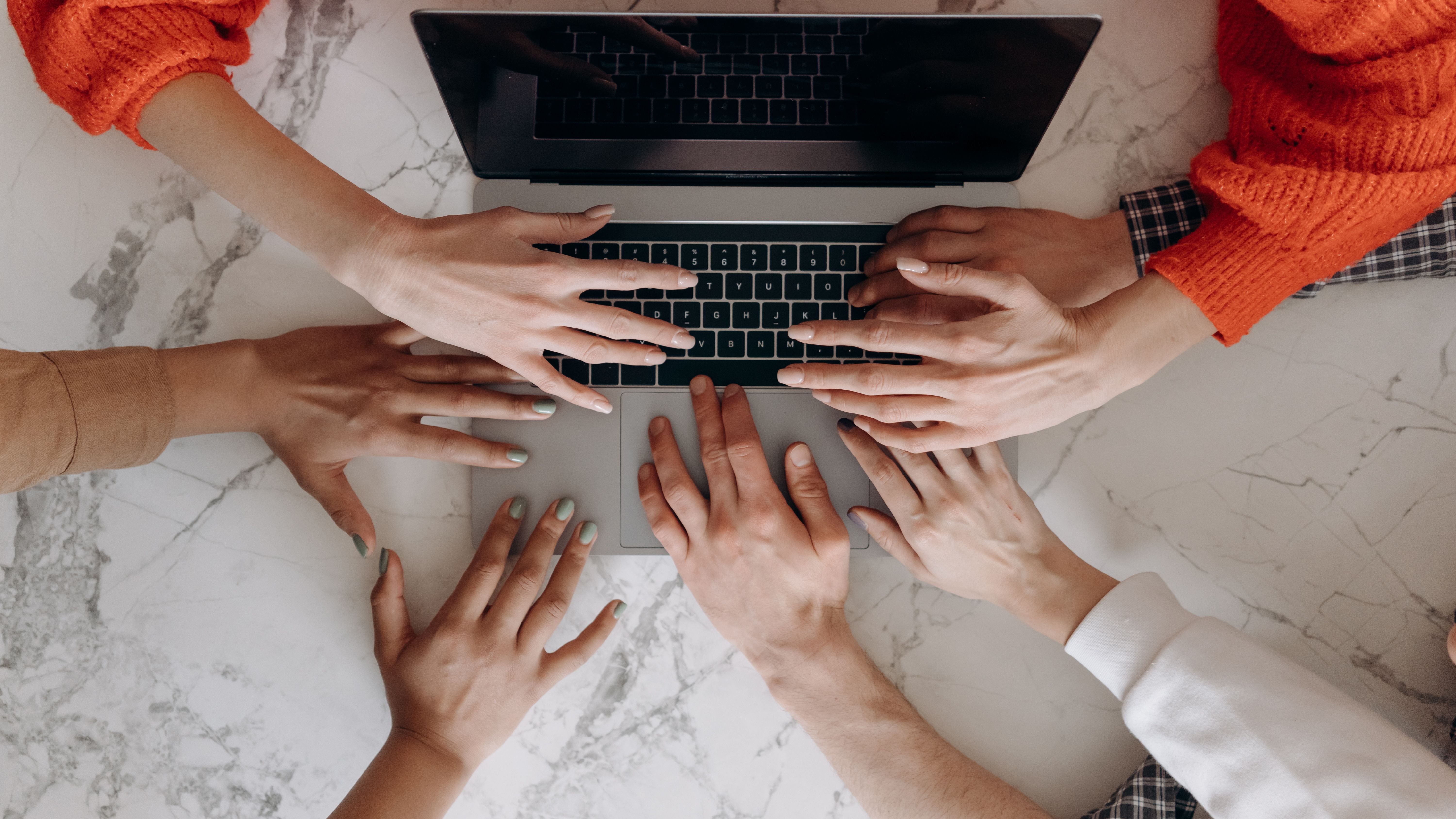 A laptop with four people's hands on the keyboard reaching in from all sides.