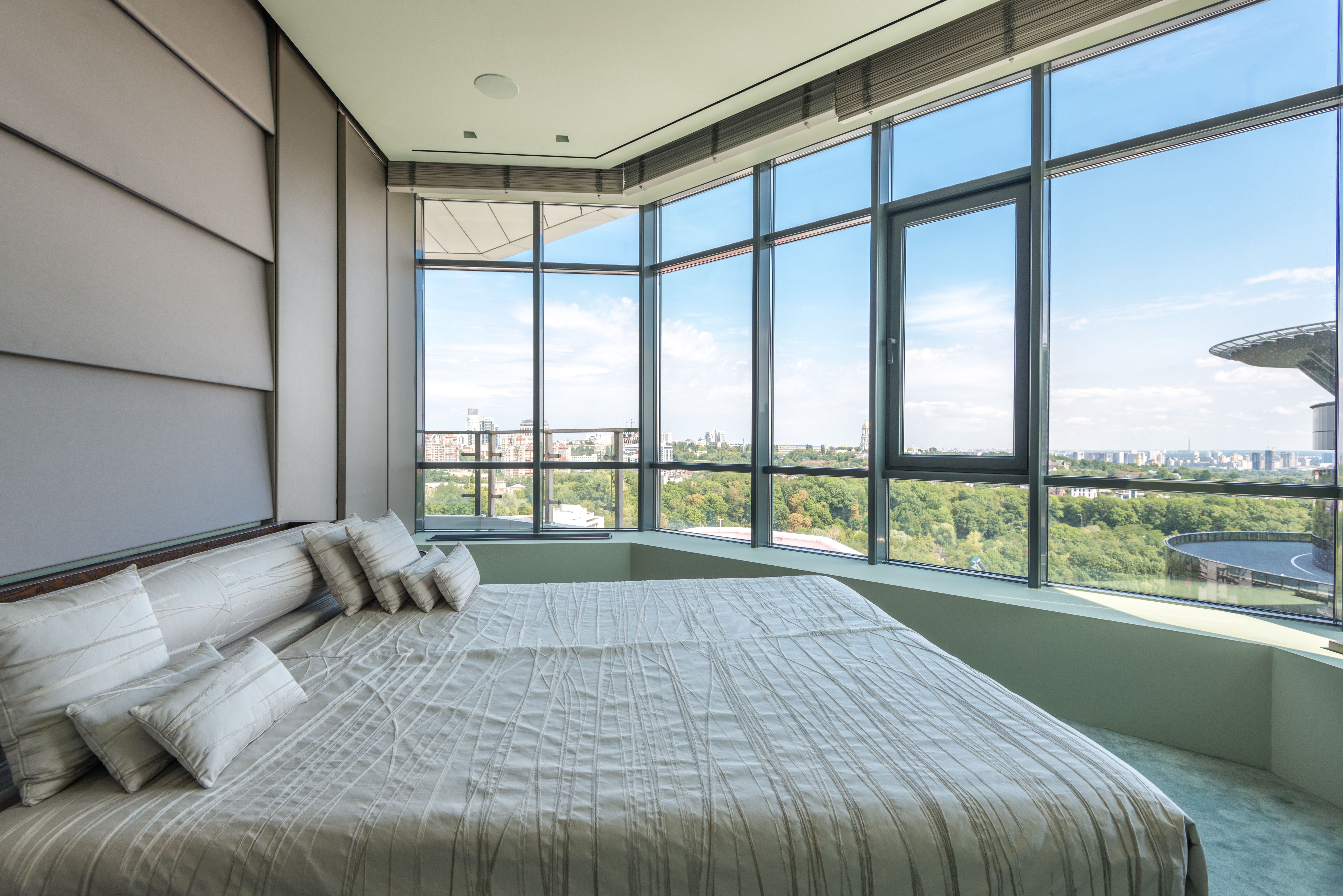 A luxury apartment bedroom with a king size bed and panoramic windows overlooking a big park in the middle of a city.